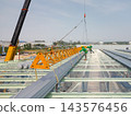 Crane lifting metal roof sheets beside workers on steel factory framing in Rayong, Thailand 143576456