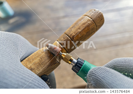Detailed close-up of a skilled craftsman wearing gray protective gloves carefully sanding and polishing a rustic handmade wooden handle using a green mini rotary tool with a conical abrasive grinding Detailed close-up of a skilled craftsman wearing gray protective gloves carefully sanding and polishing a rustic handmade wooden handle using a green mini rotary tool with a conical abrasive grinding 143576484
