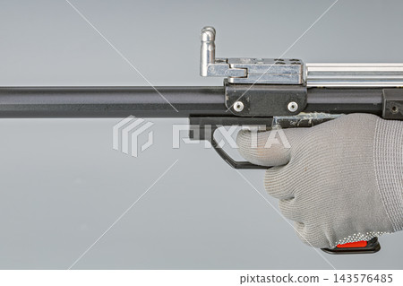 Detailed studio close-up of a hand wearing a gray textile safety glove gripping the black trigger mechanism and handle of a compact pistol crossbow immediately after shooting, shown against a plain Detailed studio close-up of a hand wearing a gray textile safety glove gripping the black trigger mechanism and handle of a compact pistol crossbow immediately after shooting, shown against a plain 143576485