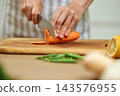 Person's hands preparing healthy food, slicing fresh carrot into thin pieces on a wooden cutting board in a kitchen 143576955