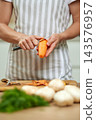 Woman's hands in a kitchen apron peeling fresh carrot with a peeler, preparing healthy vegetables on a wooden cutting board 143576957