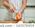 Person wearing apron peeling a raw carrot with vegetable peeler over a cutting board, preparing healthy food for cooking 143576959
