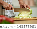 Person slicing green cabbage with knife on a wooden cutting board, preparing fresh vegetables in a kitchen 143576961