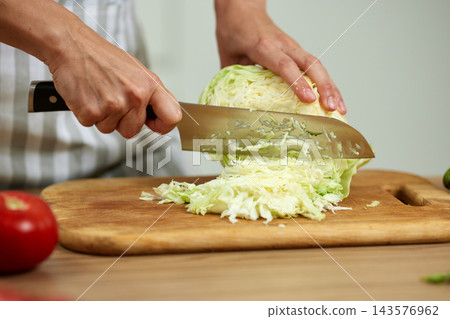 Hands cutting green cabbage with a sharp knife on a wooden cutting board, making a healthy vegetable salad 143576962