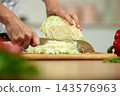 Person's hands using a knife to finely chop fresh green cabbage on a wooden board, preparing healthy food in a kitchen 143576963