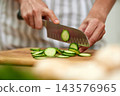 Person's hands using a sharp knife to prepare fresh organic cucumber slices on a rustic wooden cutting board, close-up 143576965