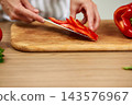 Hands slicing fresh red bell pepper into strips on a wooden cutting board, preparing healthy food in a kitchen environment 143576967