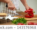 Hands in apron uniform cutting fresh bell pepper with a chef knife on a wooden board, preparing vegetable for cooking 143576968