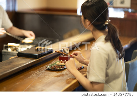 一位年輕女子在烤雞串店的櫃檯前享用烤雞串和清酒，這是她的背影。 143577793