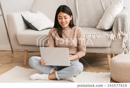 Young Asian woman networking on laptop, sitting on floor near sofa at home Young Asian woman networking on laptop, sitting on floor near sofa at home 143577819