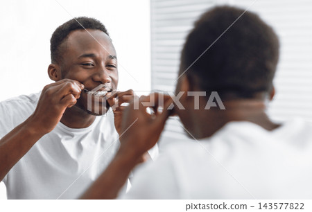 Toothy African Man Flossing And Cleaning Teeth Using Tooth Floss Standing Near Mirror In Modern Bathroom At Home In The Morning. Toothcare And Healthy Oral Hygiene Concept. Selective Focus 143577822