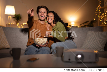 Joyful Couple Watching Film Online Using Home Cinema Projector Enjoying Evening Together On Weekend Sitting On Sofa In Living Room Indoors. Family Leisure, Entertainment And Fun Concept 143577848