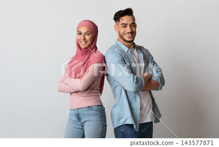 Portrait of confident happy islamic couple standing back to back with folded arms, positive arab man and muslim woman in hijab posing together on grey studio background, looking and smiling at camera Portrait of confident happy islamic couple standing back to back with folded arms, positive arab man and muslim woman in hijab posing together on grey studio background, looking and smiling at camera 143577871