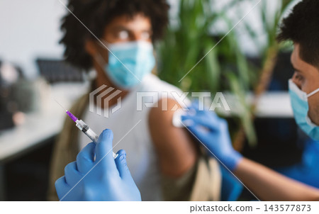 Clinical trial for new modern vaccine during global pandemic. Guy in medical face mask getting injection at hospital. Doctor preparing syringe to give a shot, rubbing arm with cotton pad Clinical trial for new modern vaccine during global pandemic. Guy in medical face mask getting injection at hospital. Doctor preparing syringe to give a shot, rubbing arm with cotton pad 143577873