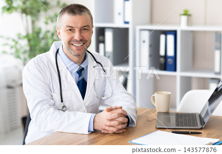 Healthcare And Medicine Concept. Portrait of cheerful mature doctor posing, smiling at camera, sitting at desk in office Healthcare And Medicine Concept. Portrait of cheerful mature doctor posing, smiling at camera, sitting at desk in office 143577876