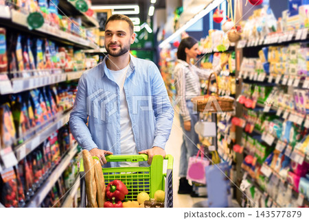 Everyday Shopping. Portrait Of Young Bearded Guy Walking With Trolley Cart Between The Aisles In Wholesale Store. Casual Man Buying Healthy Food In Shop, Looking At Shelves, Blurred Background Everyday Shopping. Portrait Of Young Bearded Guy Walking With Trolley Cart Between The Aisles In Wholesale Store. Casual Man Buying Healthy Food In Shop, Looking At Shelves, Blurred Background 143577879