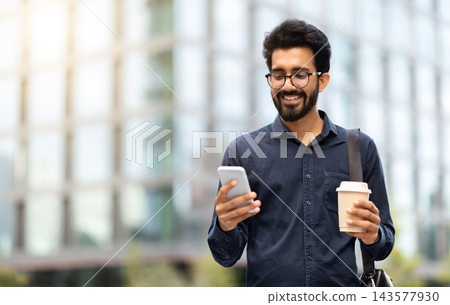 Cheerful millennial indian guy employee going to office in the morning, using phone on the street, drinking takeaway coffee. Happy young eastern businessman checking email on smartphone, copy space Cheerful millennial indian guy employee going to office in the morning, using phone on the street, drinking takeaway coffee. Happy young eastern businessman checking email on smartphone, copy space 143577930