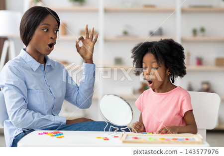 Pretty little girl attending speech therapy session at clinic, looking at mirror, beautiful young african american woman speech-language pathologist working on sounds with kid, articulating Pretty little girl attending speech therapy session at clinic, looking at mirror, beautiful young african american woman speech-language pathologist working on sounds with kid, articulating 143577976