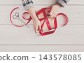 DIY hobby. Woman's hands wrapping christmas or other holiday handmade present in paper with red ribbon. Making bow at present box, decoration of gift. Scissors on white wooden table, top view. 143578085
