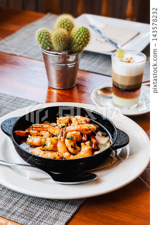 Sizzling garlic prawns on a restaurant table with coffee. Prawns sizzle in garlic and olive oil in a small cast iron dish with a barraquito coffee behind. Spanish tapas culture for restaurant marketin 143578232