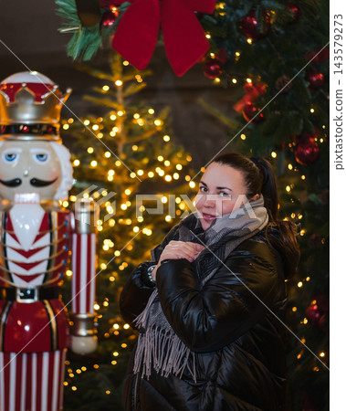 Woman in Winter Coat by Nutcracker and Christmas Lights. Young woman in a black puffer coat and scarf poses with crossed arms beside a nutcracker figure in front of a glowing Christmas tree with warm  143579273