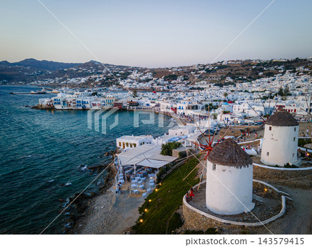 Aerial Sunset View of Mykonos Old Town and Port. A stunning aerial sunset view of Mykonos Old Town, featuring its iconic whitewashed houses, historic windmills, and vibrant port. The golden hues of th Aerial Sunset View of Mykonos Old Town and Port. A stunning aerial sunset view of Mykonos Old Town, featuring its iconic whitewashed houses, historic windmills, and vibrant port. The golden hues of th 143579415