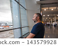 Side profile of a traveler watching the runway. A man in side profile watches activity on the runway from inside a bright airport terminal. Natural light floods the wide hall around his quiet figure. 143579928
