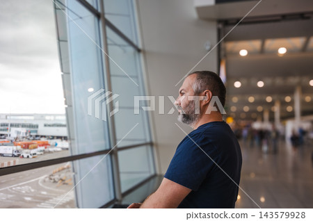 Side profile of a traveler watching the runway. A man in side profile watches activity on the runway from inside a bright airport terminal. Natural light floods the wide hall around his quiet figure. Side profile of a traveler watching the runway. A man in side profile watches activity on the runway from inside a bright airport terminal. Natural light floods the wide hall around his quiet figure. 143579928