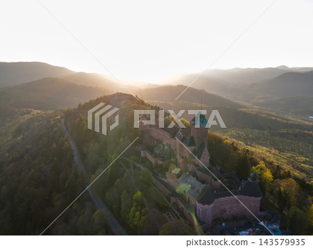 Backlit fortress glowing in sunset light. Castle Haut-Koenigsbourg, Alsace, France - April 10, 2026 - Backlit fortress glowing in sunset light, amber rim highlights towers, long access road visible Backlit fortress glowing in sunset light. Castle Haut-Koenigsbourg, Alsace, France - April 10, 2026 - Backlit fortress glowing in sunset light, amber rim highlights towers, long access road visible 143579935