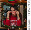 Woman Adjusting Party Glasses in Festive Christmas Frame. Playful young woman in a red sweater peers over rhinestone party sunglasses inside a handmade Christmas garland frame topped with inflatable 143579936