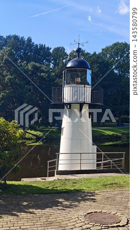 White lighthouse tower with dark domed lantern room and balcony beside a tree lined pond under blue sky in a landscaped park 143580799