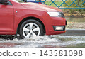 A red car navigates through a city street filled with water during a rainstorm. Puddles form around the vehicle as it moves 143581098