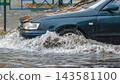 A car drives through a city street that is flooded due to heavy rain. Water splashes around the tires as it moves through the deep puddles 143581100
