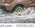 A car navigates through a flooded city street as rain pours down. Water splashes around the wheels, creating puddles and challenges for drivers 143581101