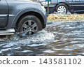 A car navigates a flooded city street as rain pours down, creating puddles and causing difficulties for vehicles in the area 143581102