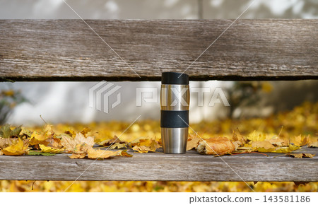 A stainless steel thermos and fresh pastry rest on a wooden bench covered with autumn leaves. The scene represents comfort and peaceful recreation. A stainless steel thermos and fresh pastry rest on a wooden bench covered with autumn leaves. The scene represents comfort and peaceful recreation. 143581186