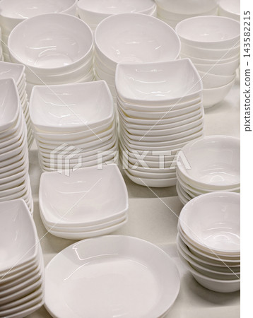 Ceramic white tableware arranged in stacked sets including plates saucers and salad bowls displayed on retail market shelves creating a clean and simple kitchenware assortment for everyday use Ceramic white tableware arranged in stacked sets including plates saucers and salad bowls displayed on retail market shelves creating a clean and simple kitchenware assortment for everyday use 143582215