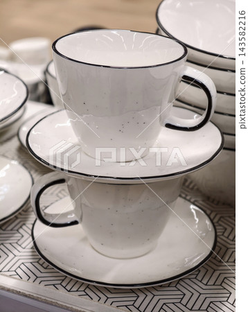 Two ceramic cups on saucers stacked neatly creating a minimal kitchenware composition with clean white finish and practical tableware concept for modern store shelf display Two ceramic cups on saucers stacked neatly creating a minimal kitchenware composition with clean white finish and practical tableware concept for modern store shelf display 143582216