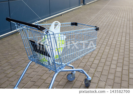 Light blue metal shopping cart carrying single plastic grocery bag stands on paved ground outside supermarket wall. Retail trade, consumerism, purchasing power, and grocery store service Light blue metal shopping cart carrying single plastic grocery bag stands on paved ground outside supermarket wall. Retail trade, consumerism, purchasing power, and grocery store service 143582700