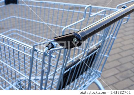 Empty metal shopping trolley stands outdoors near supermarket pavement. Economic crisis, inflation impact, reduced purchasing power, and modern consumerism concept Empty metal shopping trolley stands outdoors near supermarket pavement. Economic crisis, inflation impact, reduced purchasing power, and modern consumerism concept 143582701