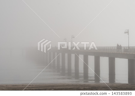 Long pier extending into heavy fog over calm sea water. Long pier with lanterns disappearing into dense white fog on a misty day, with silhouettes of people fishing, walking along bridge Long pier extending into heavy fog over calm sea water. Long pier with lanterns disappearing into dense white fog on a misty day, with silhouettes of people fishing, walking along bridge 143583893