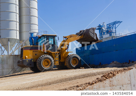 Heavy machinery transfers dirt at a construction site near a shipping dock during midday operations 143584054