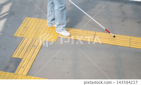A blind woman walks using a cane and tactile tiles. A blind woman walks using a cane and tactile tiles. 143584217