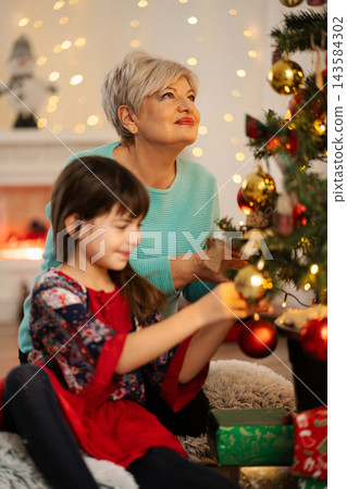 Grandchild and grandmother decorating Christmas tree during holidays Grandchild and grandmother decorating Christmas tree during holidays 143584302