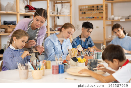 Children paint ceramic work-pieces at table with teacher in art studio 143587929