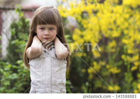 Portrait of a pretty little long-haired blond preschool girl in sleeveless white dress smiling shyly into camera against blurred outdoors background. Innocent happy childhood concept. Portrait of a pretty little long-haired blond preschool girl in sleeveless white dress smiling shyly into camera against blurred outdoors background. Innocent happy childhood concept. 143588881