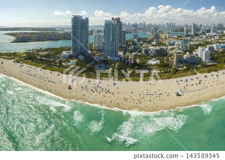 Travel destination and tourism infrastructure in southern USA. South Beach sandy surface with tourists relaxing on hot Florida sun. Miami Beach city with high luxury hotels and condos Travel destination and tourism infrastructure in southern USA. South Beach sandy surface with tourists relaxing on hot Florida sun. Miami Beach city with high luxury hotels and condos 143589345