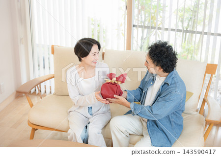一位日本丈夫在客廳的沙發上向妻子贈送禮物；妻子接過禮物時，這對恩愛的夫妻臉上露出了笑容。 143590417