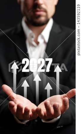 Businessman holding a sign for the year 2027 with arrows pointing upward in a professional setting Businessman holding a sign for the year 2027 with arrows pointing upward in a professional setting 143592119