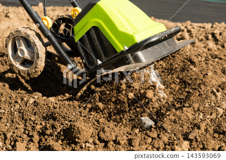 A bright green electric tiller churns through loose, brown soil in a garden bed, preparing it for planting. The cultivated earth features clumps and texture, awaiting seedlings in the spring. A bright green electric tiller churns through loose, brown soil in a garden bed, preparing it for planting. The cultivated earth features clumps and texture, awaiting seedlings in the spring. 143593069